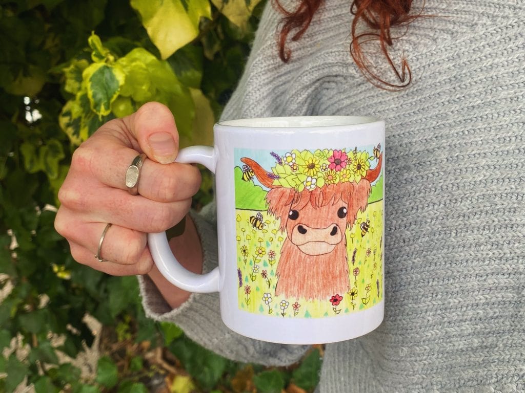 A woman's hand holding our competition winning mug with a highland cow drawing on it.
