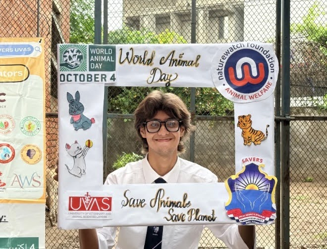 A smiling student stands behind a decorated frame promoting World Animal Day. The frame includes drawings of animals, the Naturewatch Foundation logo, and the text “World Animal Day. October 4. Save Animals, Save Planet."